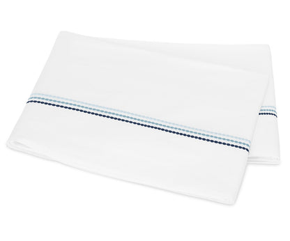 A folded white sheet from the Matouk Amara Triple Pearl Bedding Collection, featuring two parallel stripes—one light blue and one dark blue—with a dotted pattern near one edge and an elegant embroidered design in luxurious Amara sateen.