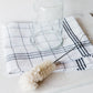 The Be Home Bistro Flute Cleaning Brush is arranged with a glass jar and checkered dish towel on a white surface, perfect for efficient glassware cleaning.