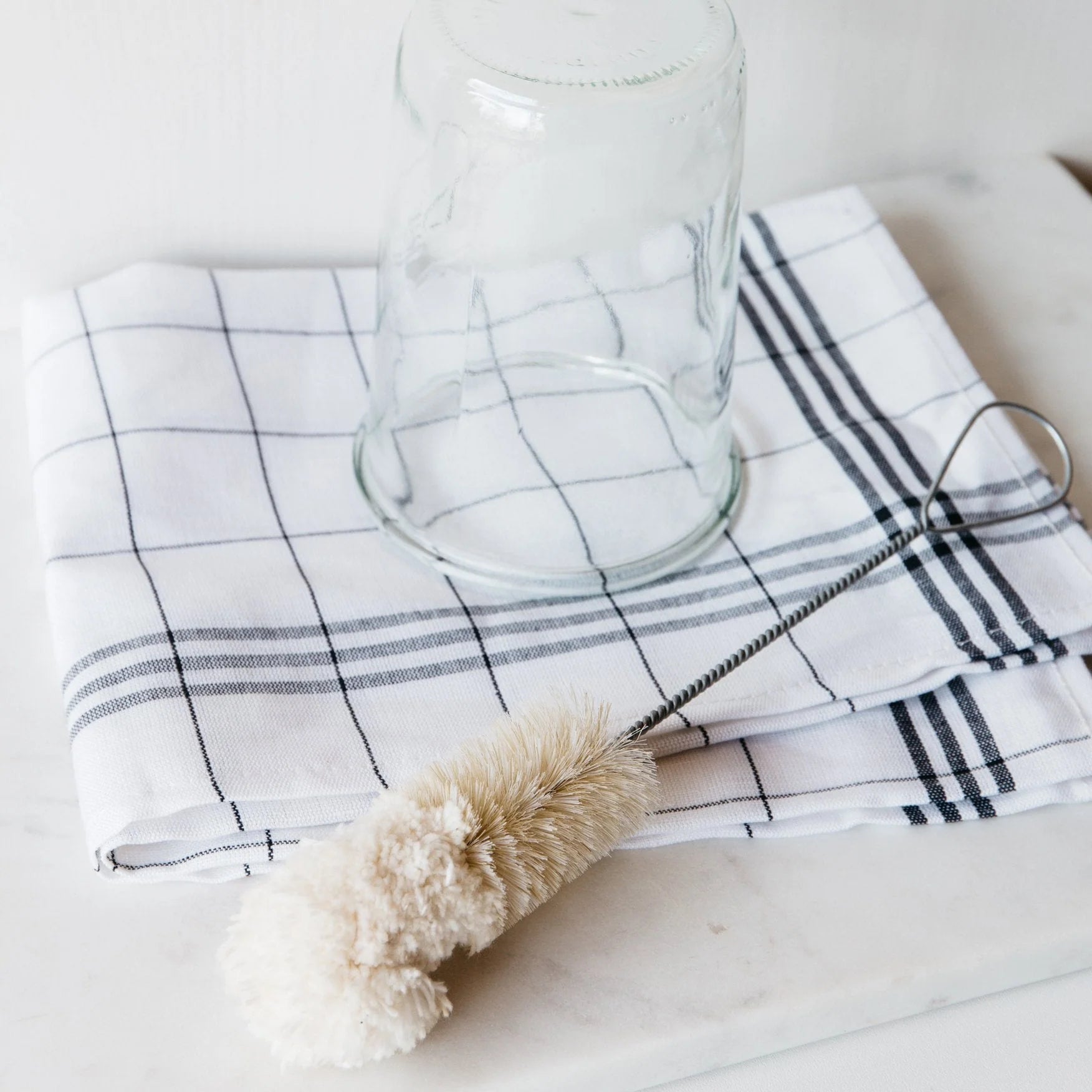The Be Home Bistro Flute Cleaning Brush is arranged with a glass jar and checkered dish towel on a white surface, perfect for efficient glassware cleaning.