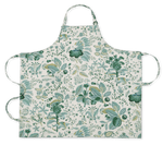 The Linen Apron Collection by Matouk features printed linen aprons with neck and waist ties, showcasing green and yellow botanical designs on a white background, inspired by Schumacher prints.