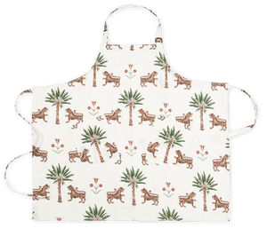 The Matouk Linen Apron Collection features a white linen apron with two front pockets, adjustable neck and waist straps, and an all-over pattern of tigers, palm trees, and red flowers inspired by Schumacher prints.