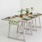 A minimalist dining table set with green and white print tableware from Pomegranate Inc. Dancing Artichokes Collection, a matching table runner, woven placemats, and hand painted globe artichokes as centerpieces. The table is placed on a white floor in a brightly lit room.