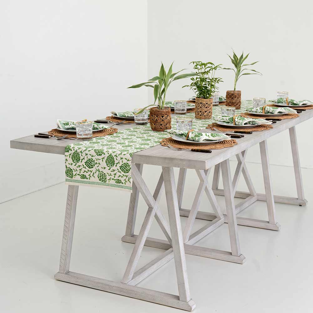 A minimalist dining table set with green and white print tableware from Pomegranate Inc. Dancing Artichokes Collection, a matching table runner, woven placemats, and hand painted globe artichokes as centerpieces. The table is placed on a white floor in a brightly lit room.