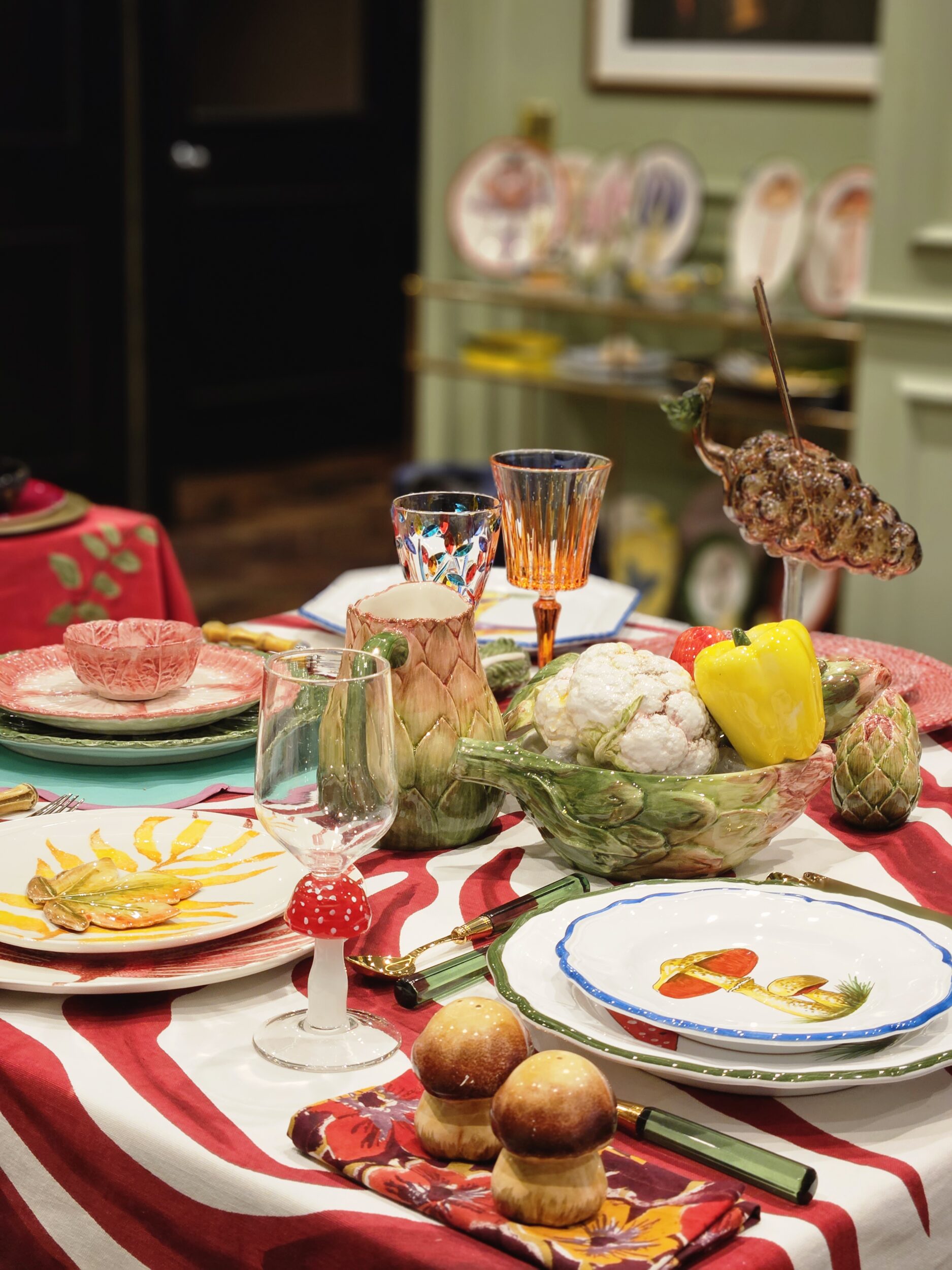 A dining table set with Les Ottomans’ Artichoke Decor and Serveware Collection, colorful plates, glassware, and vegetable-themed accents on a red and white striped tablecloth.