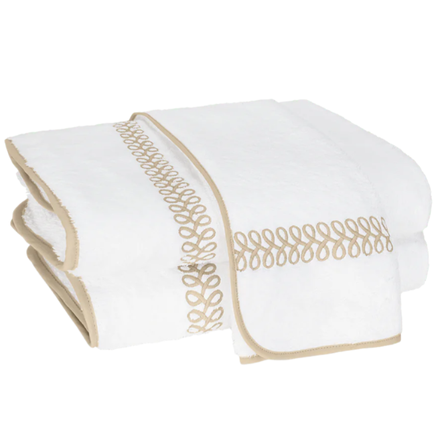 Two white towels from the Matouk Astor Braid Bath Collection, featuring beige embroidered loops and trim, are neatly folded and stacked.