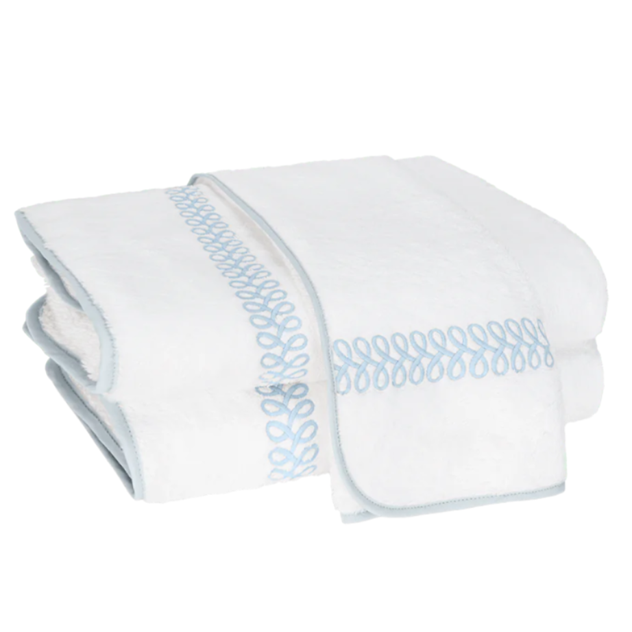 A folded towel from the Matouk Astor Braid Bath Collection, crafted of luxurious long-staple cotton with light blue embroidered Astor Braid loops and blue trim, is displayed on a white background.