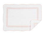The Matouk Astor Braid Bath Collection features a white rectangular blanket crafted from long-staple cotton with a subtle pink embroidered border, scalloped edges, and one corner folded to show the reverse side.