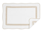 The Matouk Astor Braid Bath Collection mat features a white rectangular design with scalloped edges, a beige braid border, and plush long-staple cotton terry. A folded corner reveals the soft Cairo terry underside.