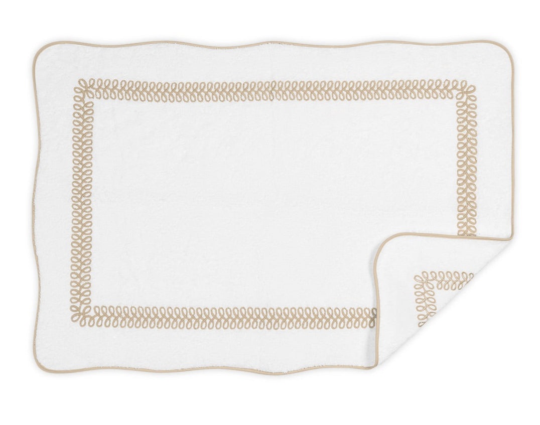 The Matouk Astor Braid Bath Collection mat features a white rectangular design with scalloped edges, a beige braid border, and plush long-staple cotton terry. A folded corner reveals the soft Cairo terry underside.