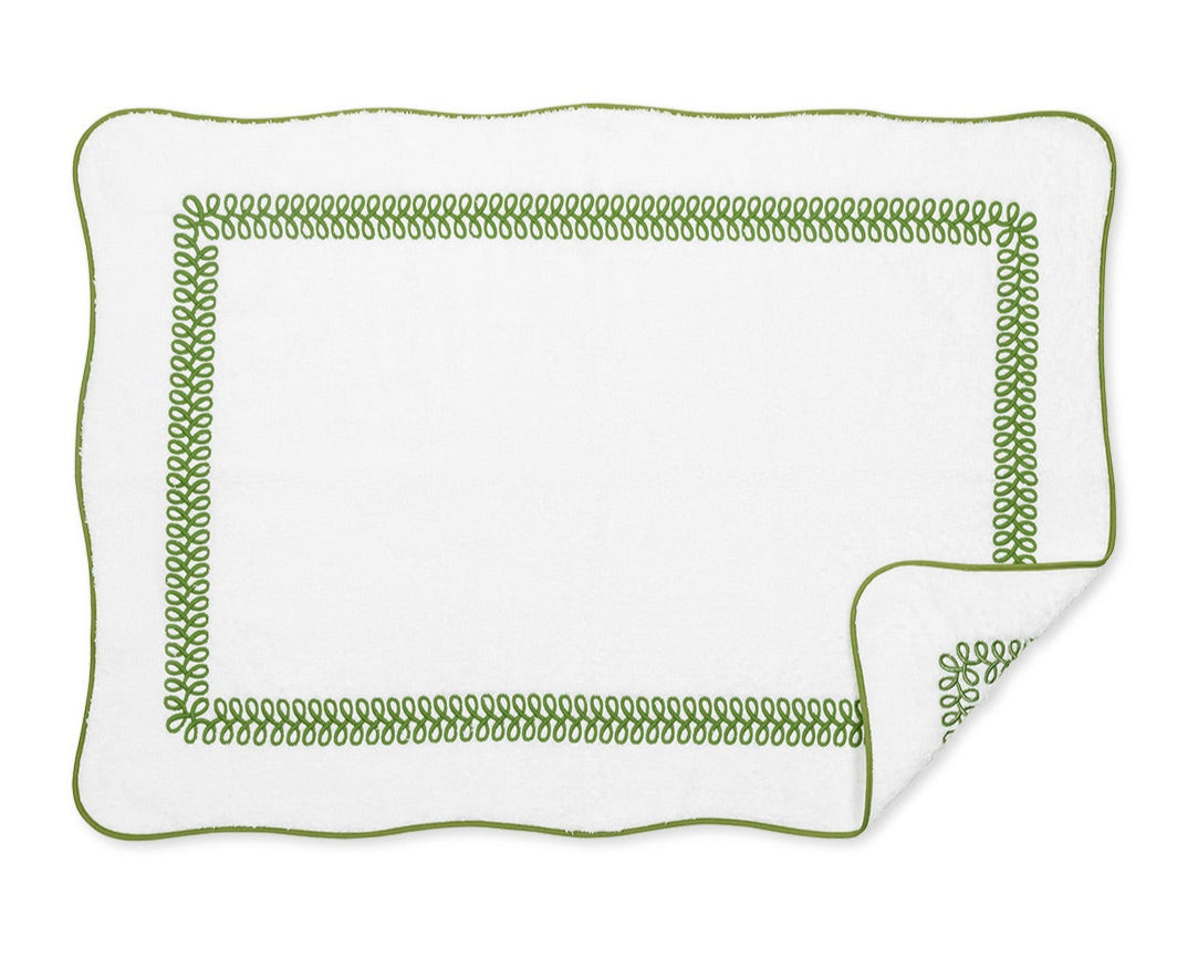 The Astor Braid Bath Collection by Matouk features a white rectangular bath mat with scalloped edges, made from long-staple cotton Cairo terry, detailed with a green looped border and a corner turned up to show the reverse pattern.