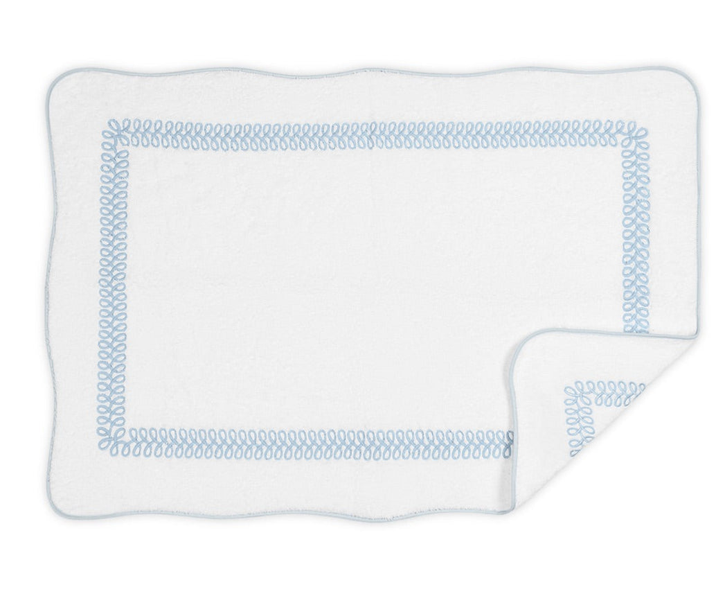 The Matouk Astor Braid Bath Collection features a white rectangular blanket made of long-staple cotton with a blue braid border, scalloped edges, and a folded corner showing the Cairo terry reverse.
