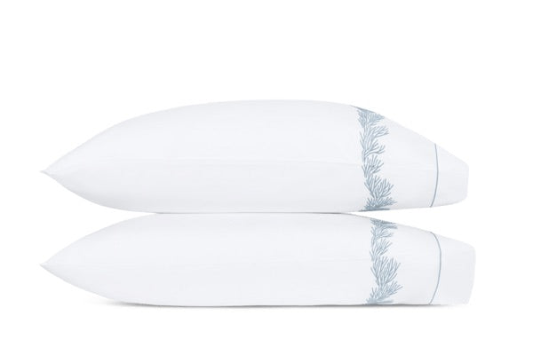 Two white pillows from the Matouk Atoll Bedding Collection, featuring an elegant blue embroidered leaf pattern, are stacked atop one another against a plain white background. Expertly crafted from soft CEYLON cotton percale with a 520 thread count, these pillows offer luxurious comfort and sophisticated style.