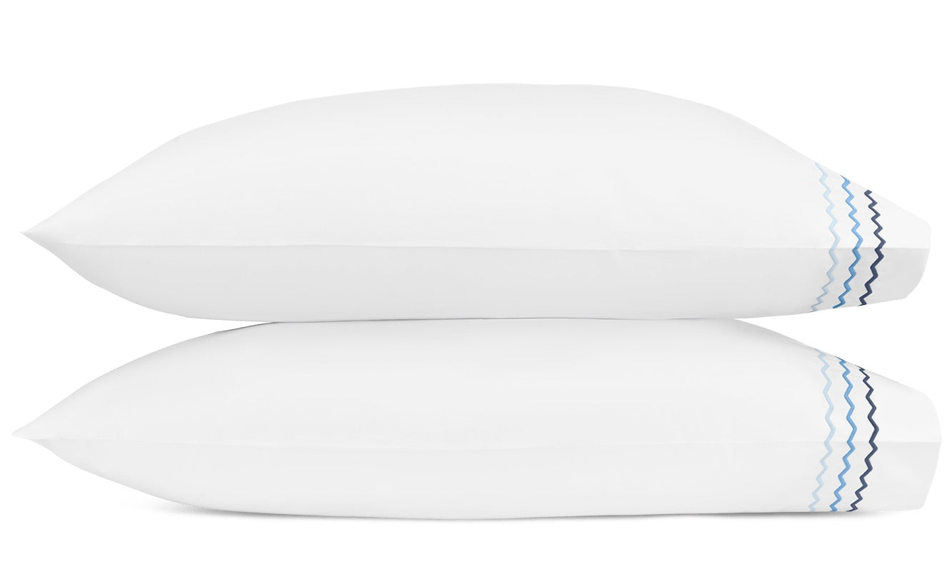 Two white pillows from the Axel Bedding Collection by Matouk, each crafted from soft long-staple cotton and featuring light blue and dark blue wavy lines embroidered in a zigzag pattern on one end, are stacked against a white background.