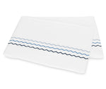 Folded white bedsheet from the Axel Bedding Collection by Matouk, featuring two zigzag embroidered lines—one light blue and one dark blue—near the edge. Crafted from long-staple cotton, this sheet adds a refined touch to any modern bed with its elegant embroidery detailing.