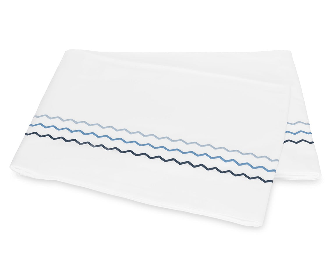 Folded white bedsheet from the Axel Bedding Collection by Matouk, featuring two zigzag embroidered lines—one light blue and one dark blue—near the edge. Crafted from long-staple cotton, this sheet adds a refined touch to any modern bed with its elegant embroidery detailing.