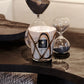 A Baobab Collection Stones Marble Candle in a decorative holder sits on a dark wooden table next to two hand-blown glass hourglasses filled with black sand.