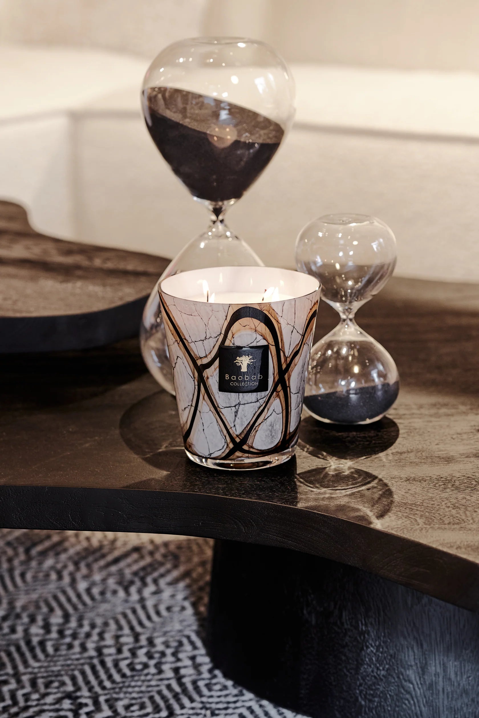 A Baobab Collection Stones Marble Candle in a decorative holder sits on a dark wooden table next to two hand-blown glass hourglasses filled with black sand.