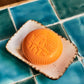 A round, orange Baobab Collection Saint Tropez soap with embossed patterns sits on a ceramic dish atop blue and teal tiles, evoking a fresh fragrance.