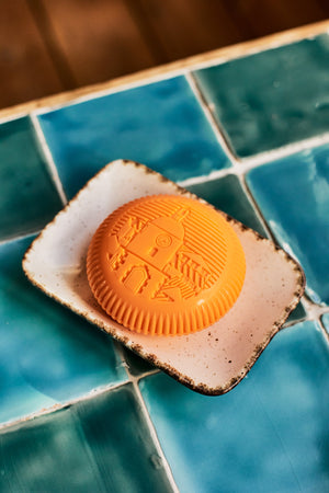 A round, orange Baobab Collection Saint Tropez soap with embossed patterns sits on a ceramic dish atop blue and teal tiles, evoking a fresh fragrance.