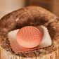 The Baobab Collection Women Body Wellness Collection coral-colored, textured soap with a delicate floral scent sits on a white ceramic dish atop a wooden surface.