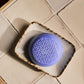 A round, purple, textured soap from the Baobab Collection Gentlemen Body Wellness line rests in a small, speckled dish on a tiled countertop, subtly evoking its refined masculine scent.