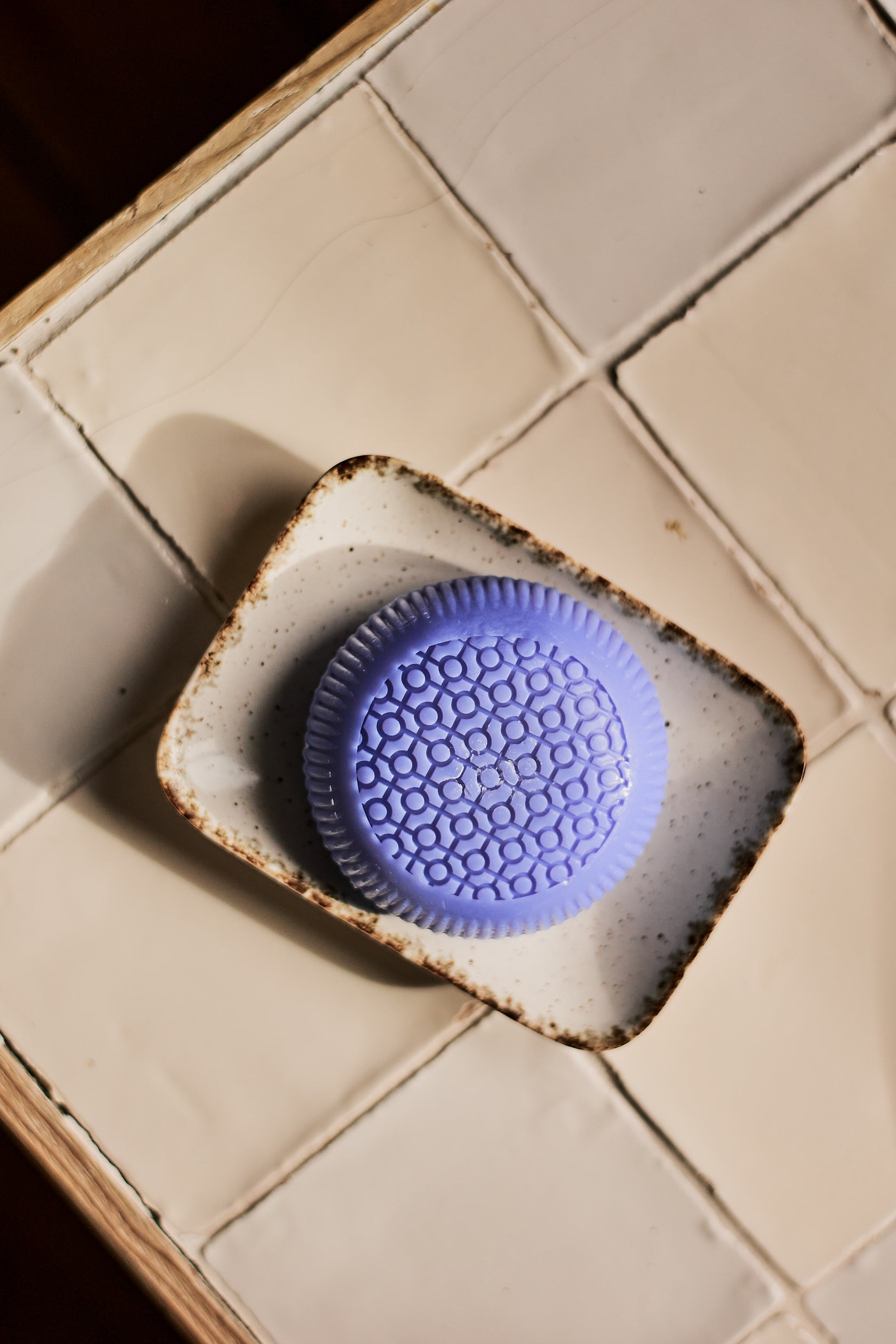 A round, purple, textured soap from the Baobab Collection Gentlemen Body Wellness line rests in a small, speckled dish on a tiled countertop, subtly evoking its refined masculine scent.