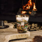 A multi-wick Baobab Collection Far West Candle in a glass holder decorated with a landscape of mountains, a ranch, and horses, sitting on a wooden table in front of a fireplace.