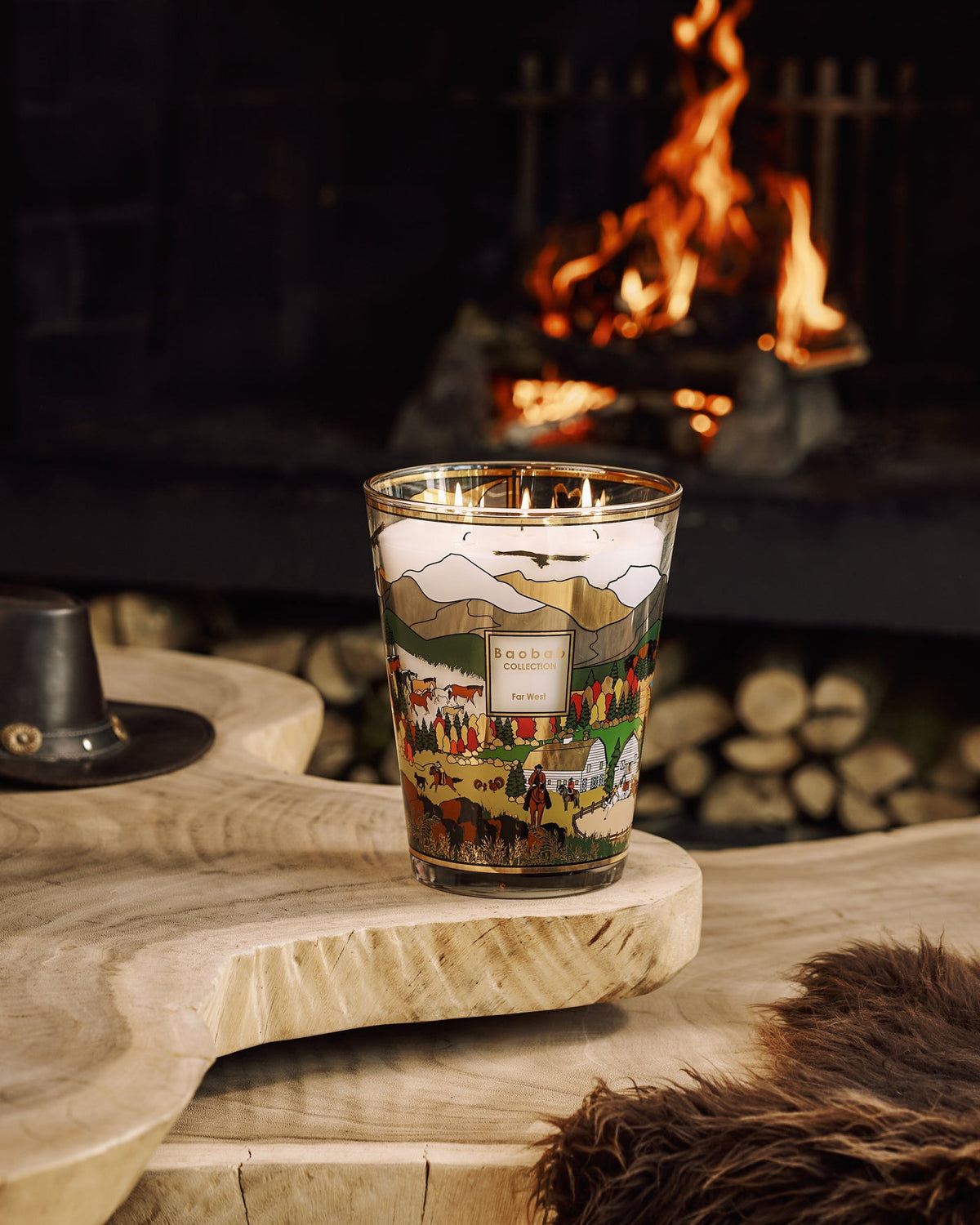 A multi-wick Baobab Collection Far West Candle in a glass holder decorated with a landscape of mountains, a ranch, and horses, sitting on a wooden table in front of a fireplace.