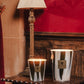 Two Baobab Collection Woods Sherwood candles in reflective holders glow on a wooden side table by a lamp, set against a red patterned wall and dark wood paneling for a cozy English countryside feel.