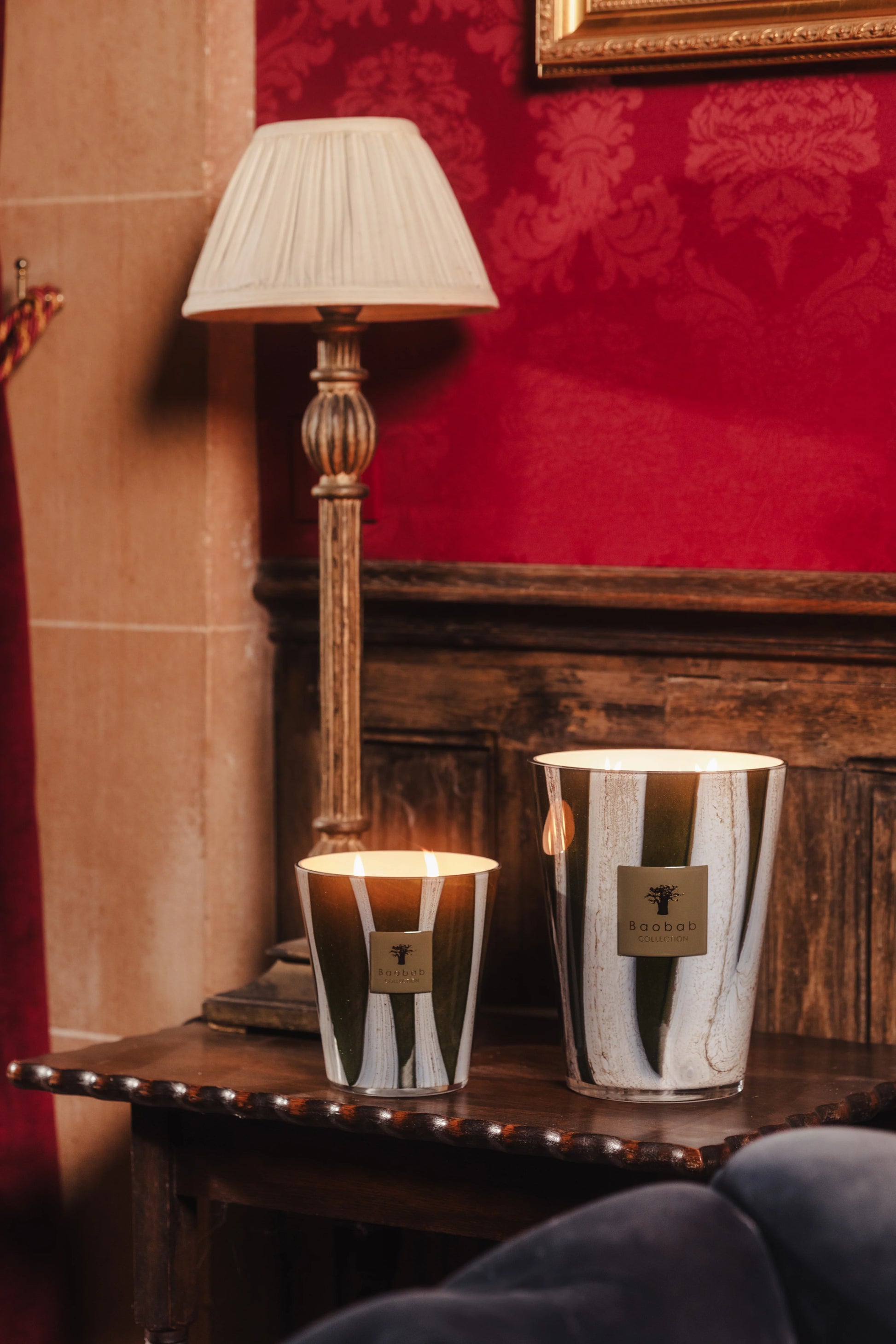 Two Baobab Collection Woods Sherwood candles in reflective holders glow on a wooden side table by a lamp, set against a red patterned wall and dark wood paneling for a cozy English countryside feel.