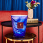 The luxurious ambiance is enhanced by a Baobab Collection Atlas Majorelle Candle resting on a gold stand in front of a plush red couch and black round table. The air is filled with the rich scent of patchouli, while blue curtains softly drape in the background.