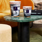 Two Baobab Collection Atlas Toubkal candles, featuring a marble pattern and the fragrance of Atlas cedar, are elegantly displayed on a green marble table beside a luxury brand book. In the background, a mustard yellow sofa beautifully complements the beige ottoman.