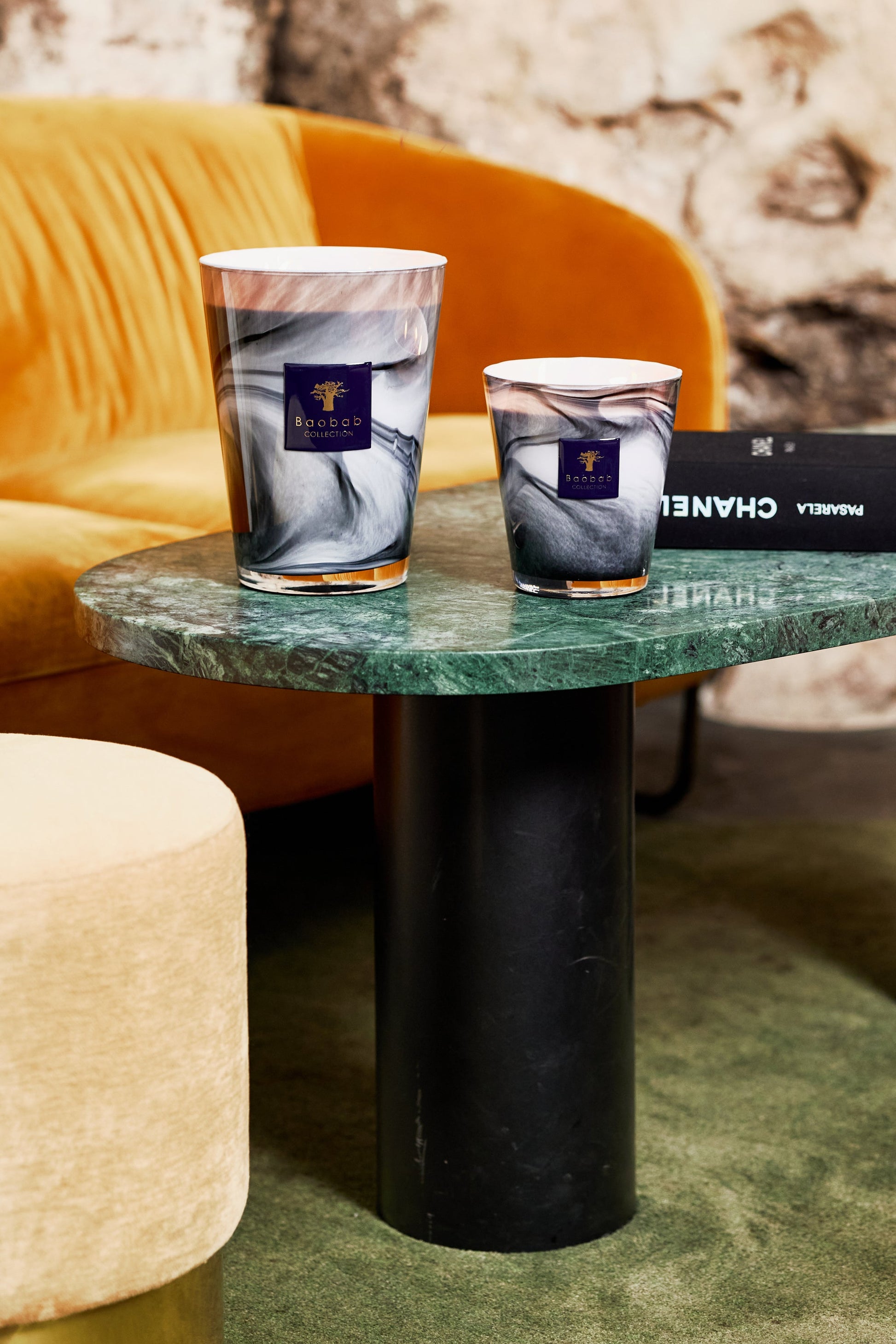 Two Baobab Collection Atlas Toubkal candles, featuring a marble pattern and the fragrance of Atlas cedar, are elegantly displayed on a green marble table beside a luxury brand book. In the background, a mustard yellow sofa beautifully complements the beige ottoman.