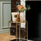 A small round table with a glass top and gold frame holds a lit Baobab Collection Atlas Tichka Candle and a vase of flowers in front of a decorative fireplace.