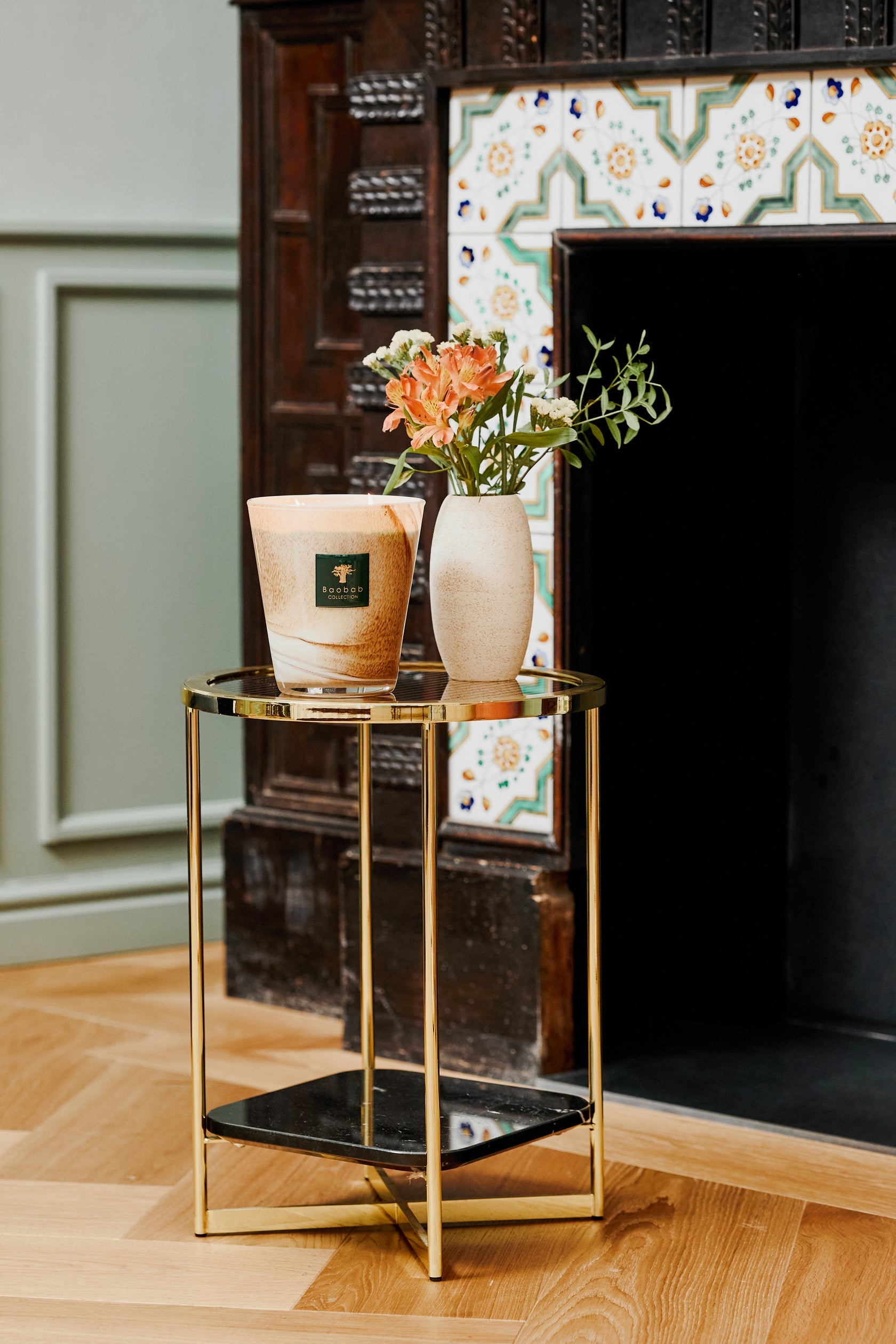 A small round table with a glass top and gold frame holds a lit Baobab Collection Atlas Tichka Candle and a vase of flowers in front of a decorative fireplace.