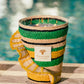 A woven candle holder with green, yellow, and natural stripes from Baobab's "Fety Milalao Candle" sits on a wooden surface near a pool. Flames flicker inside, releasing vetiver and ylang-ylang scents.