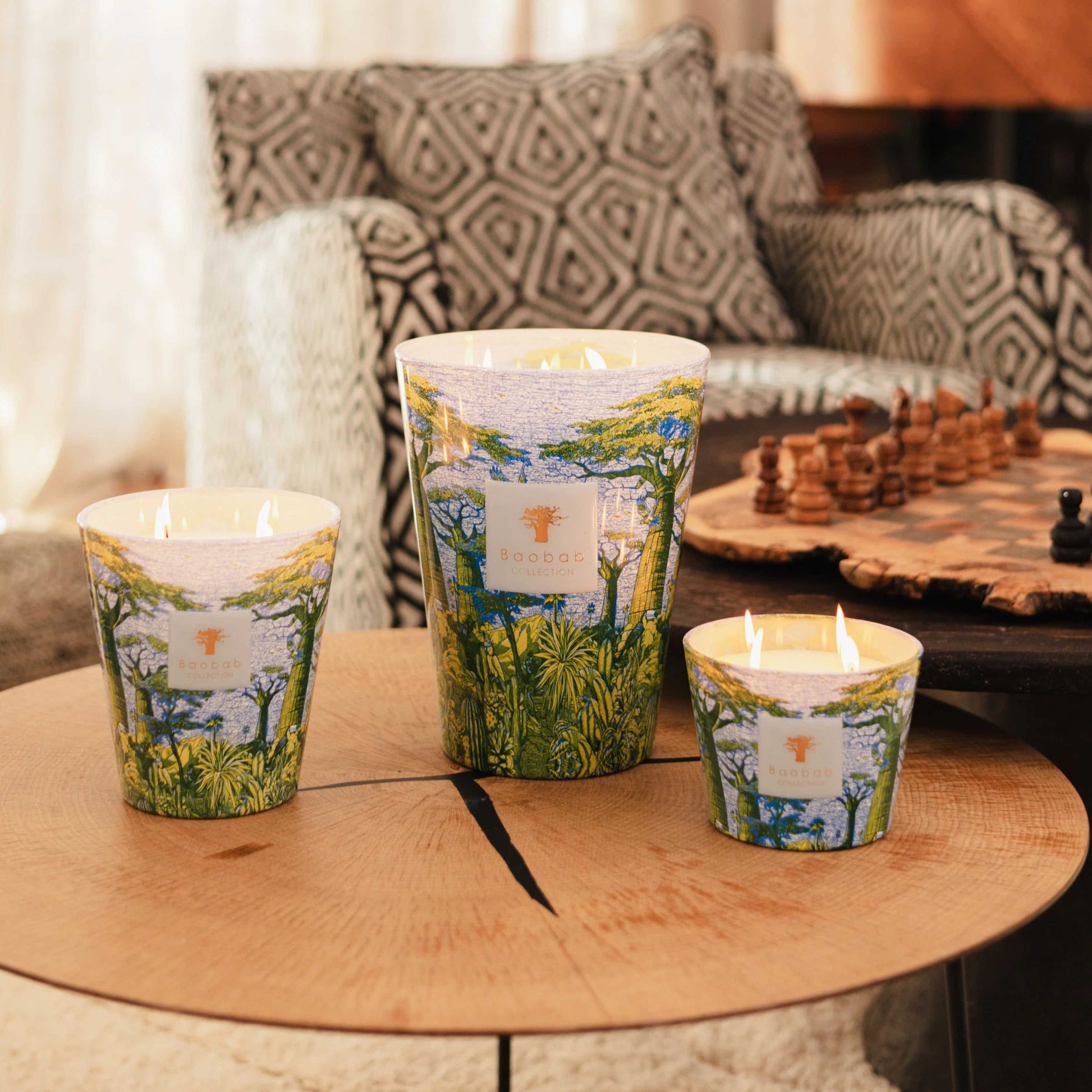 Three Baobab Collection Sacred Trees Summer Yarani patterned candles of different sizes rest on a round wooden table. A chessboard is set against patterned furniture, creating the mystique of a baobab forest.
