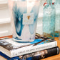 A lit Baobab Collection Riviera Leopolda Candle sits atop two stacked books—"Great Escapes Mediterranean" and "Paris"—with blue glass bottles reminiscent of the French Riviera in the background on a wooden surface.