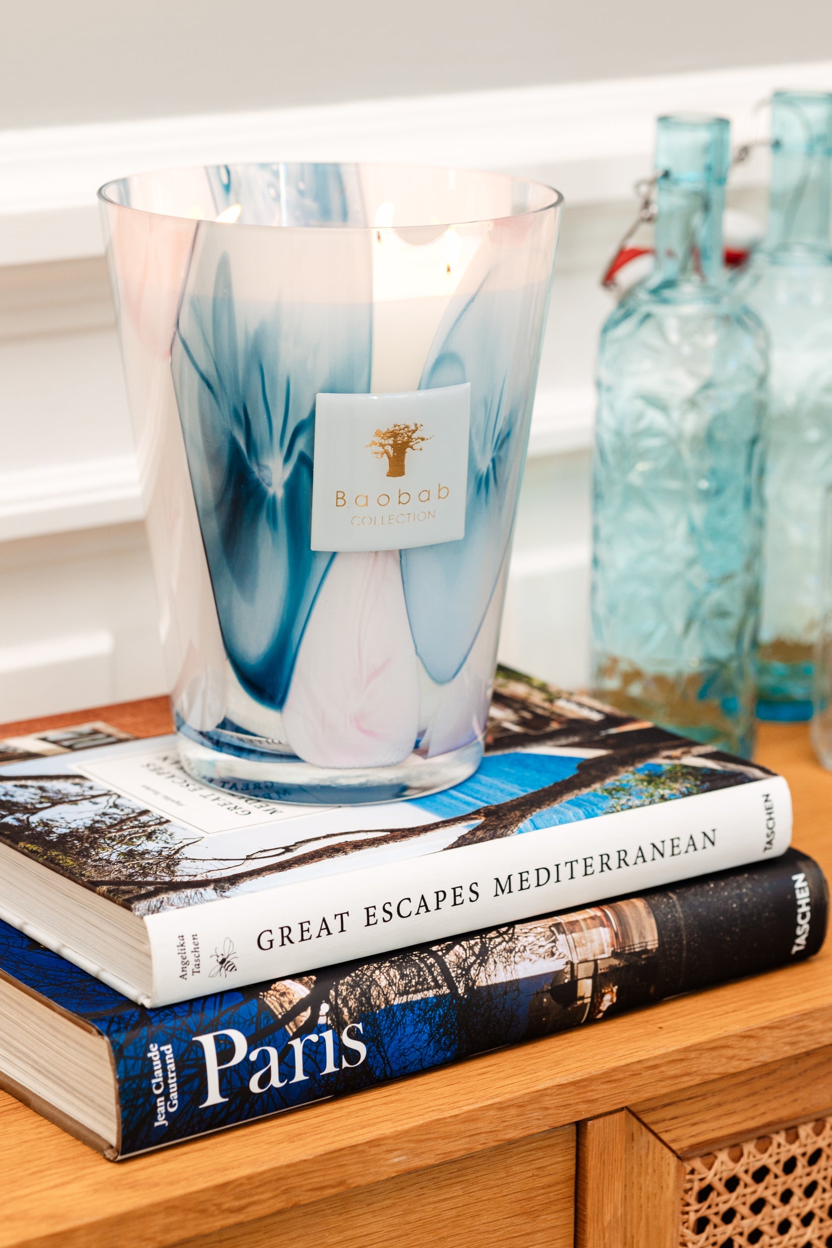 A lit Baobab Collection Riviera Leopolda Candle sits atop two stacked books—"Great Escapes Mediterranean" and "Paris"—with blue glass bottles reminiscent of the French Riviera in the background on a wooden surface.