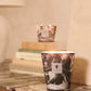 A lit Baobab Collection Leaves Manon Candle by Baobab, in a marbled hand-blown glass holder, sits on a marble-topped wooden table, with another candle and stacked books in the background.