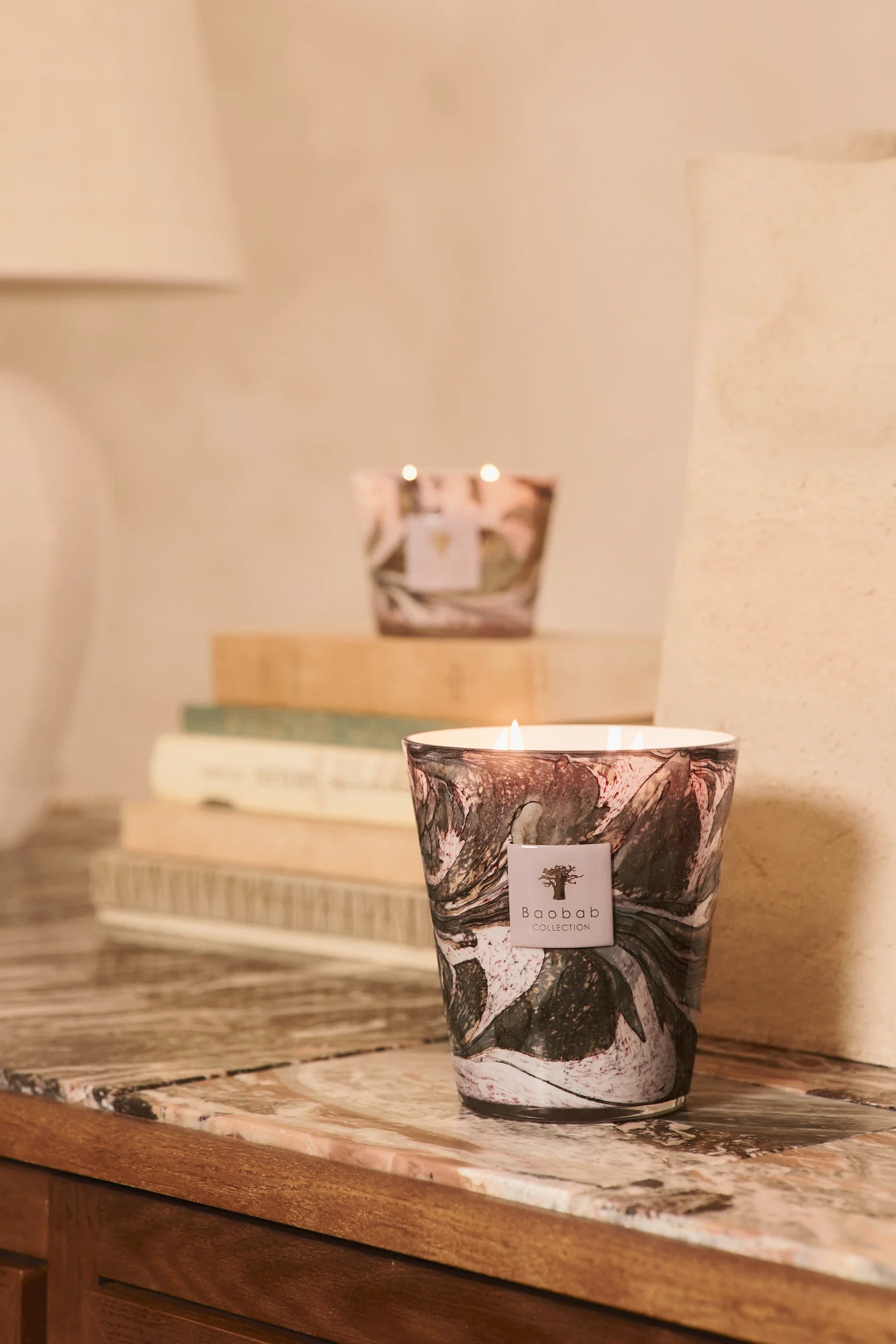 A lit Baobab Collection Leaves Manon Candle by Baobab, in a marbled hand-blown glass holder, sits on a marble-topped wooden table, with another candle and stacked books in the background.