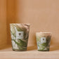 Two Baobab Leaves Marius candles by Baobab in green and white hand-blown glass sit on a beige surface, set against a peach background, capturing the charm of Provence.
