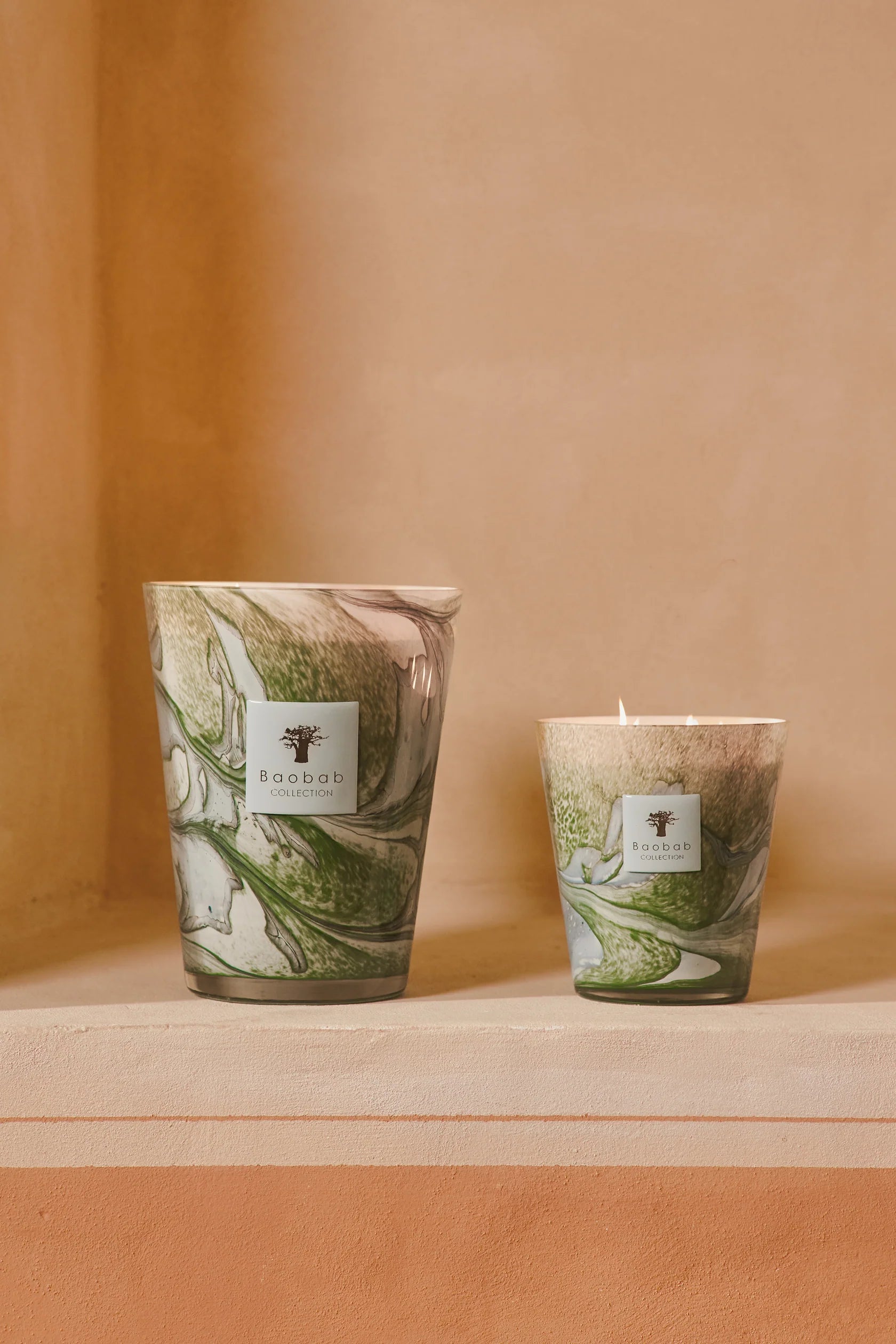Two Baobab Leaves Marius candles by Baobab in green and white hand-blown glass sit on a beige surface, set against a peach background, capturing the charm of Provence.
