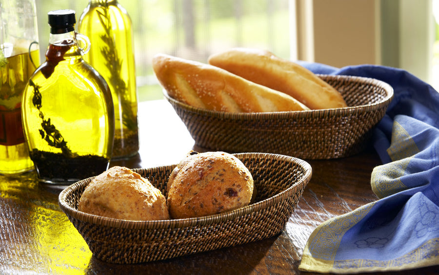 Calaisio's Oval Bread Basket Collection holds an array of fresh bread rolls and baguettes on a wooden table, accompanied by bottles of olive oil and a folded blue napkin.