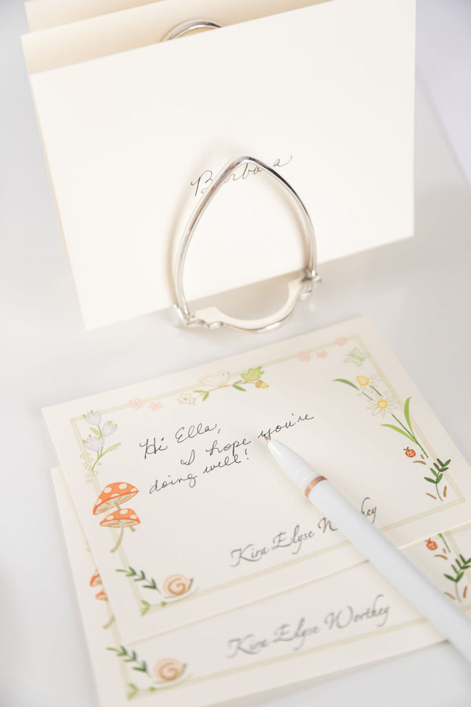 Karen Adams Letter Holder in silver plate holds stacked floral stationery cards with a handwritten note, a white pen, and an envelope—an elegant desk accessory displayed on a white surface.