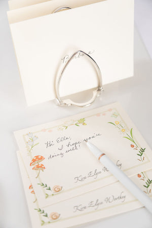 Karen Adams Letter Holder in silver plate holds stacked floral stationery cards with a handwritten note, a white pen, and an envelope—an elegant desk accessory displayed on a white surface.