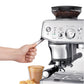 A hand uses the Breville Barista Express Impress Espresso Machine to prepare a specialty latte in a glass cup with ground coffee; pastries sit beside the cup on the tray.