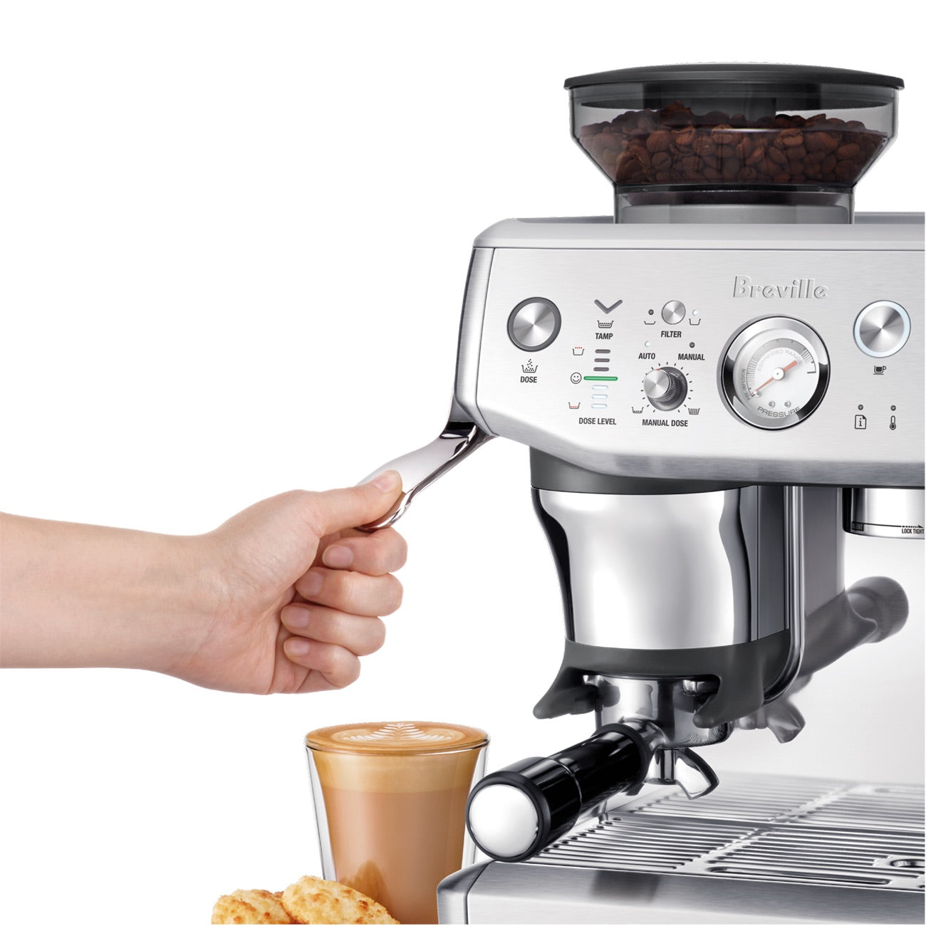 A hand uses the Breville Barista Express Impress Espresso Machine to prepare a specialty latte in a glass cup with ground coffee; pastries sit beside the cup on the tray.