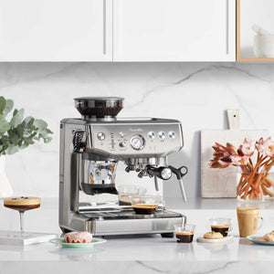 A Breville Barista Express Impress Espresso Machine sits on the kitchen counter, surrounded by specialty coffee cups, tempting desserts, and decorative plants.