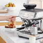 A person uses the steam wand of the Breville Barista Touch Impress Espresso Machine to make silky microfoam milk; a coffee cup, milk jug, and toast on the counter evoke a true morning barista ritual.